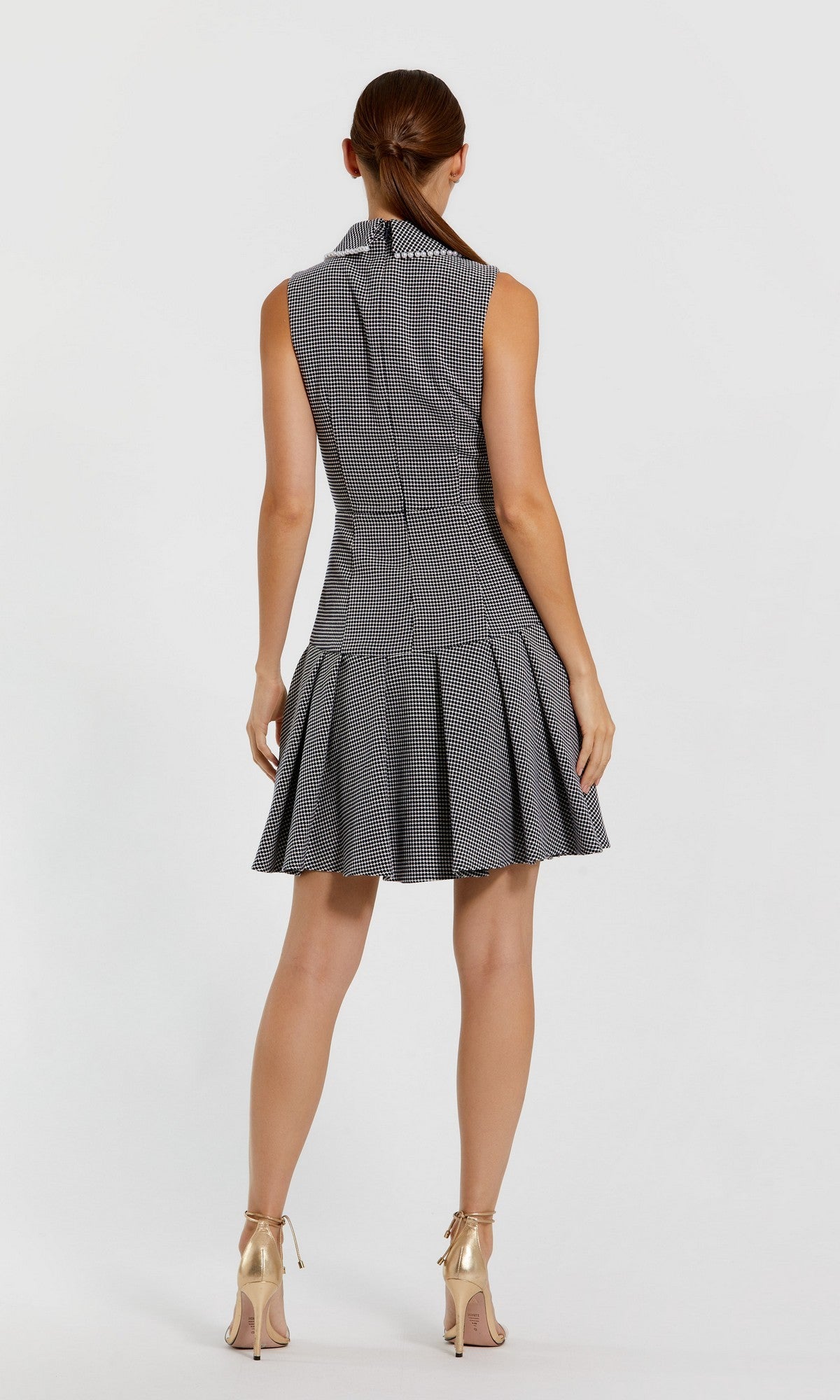 A woman stands facing away, wearing the Mac Duggal 27795 short wedding guest dress—a sleeveless, collared, gray boucle plaid mini dress with a pleated skirt—paired with beige high-heeled shoes against a plain white background.
